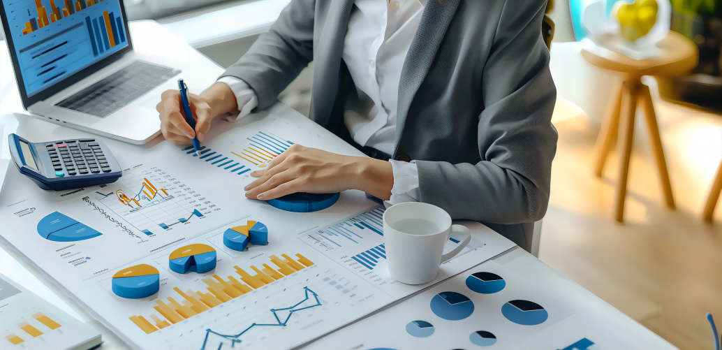 A person in business attire analyzes numerous financial charts and reports spread on a desk next to a laptop and calculator